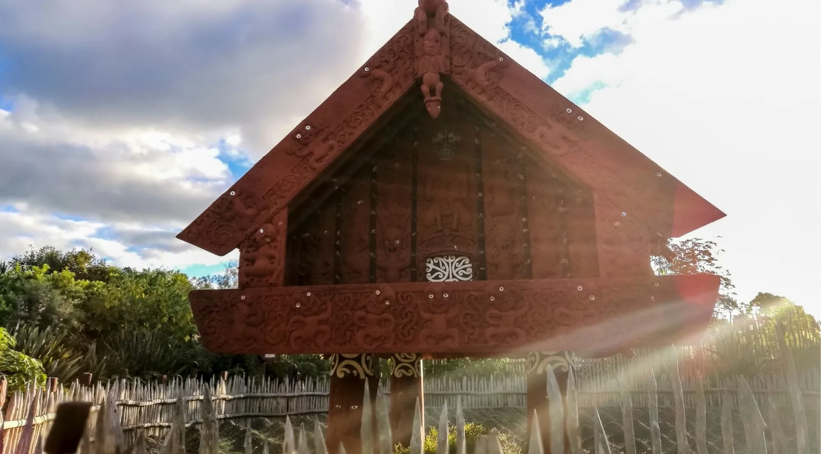 Carved pātaka storehouse raised on posts above a wooden palisade, backlit by low sun