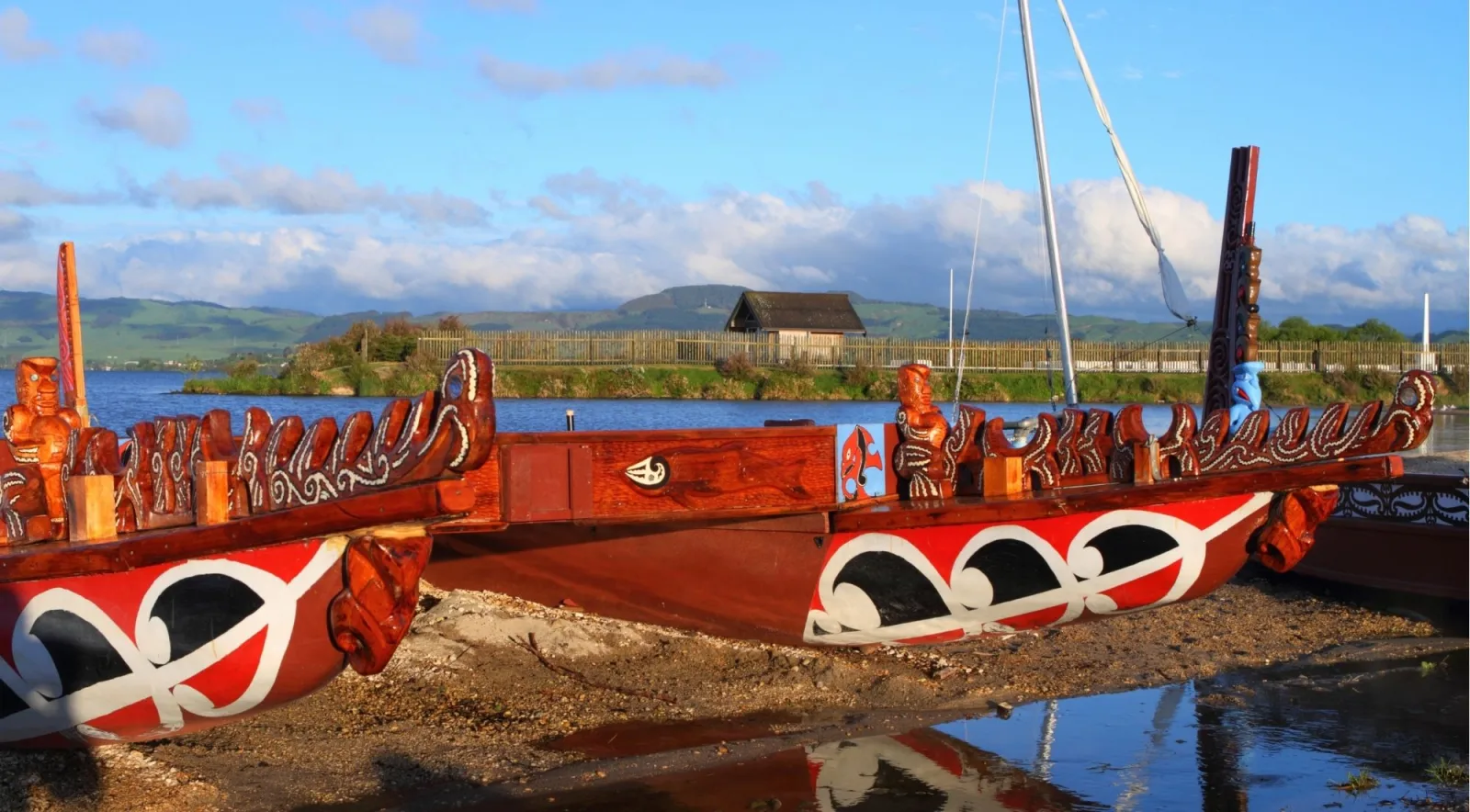 Two carved waka taua drawn up on the shore of Lake Rotorua under a blue sky