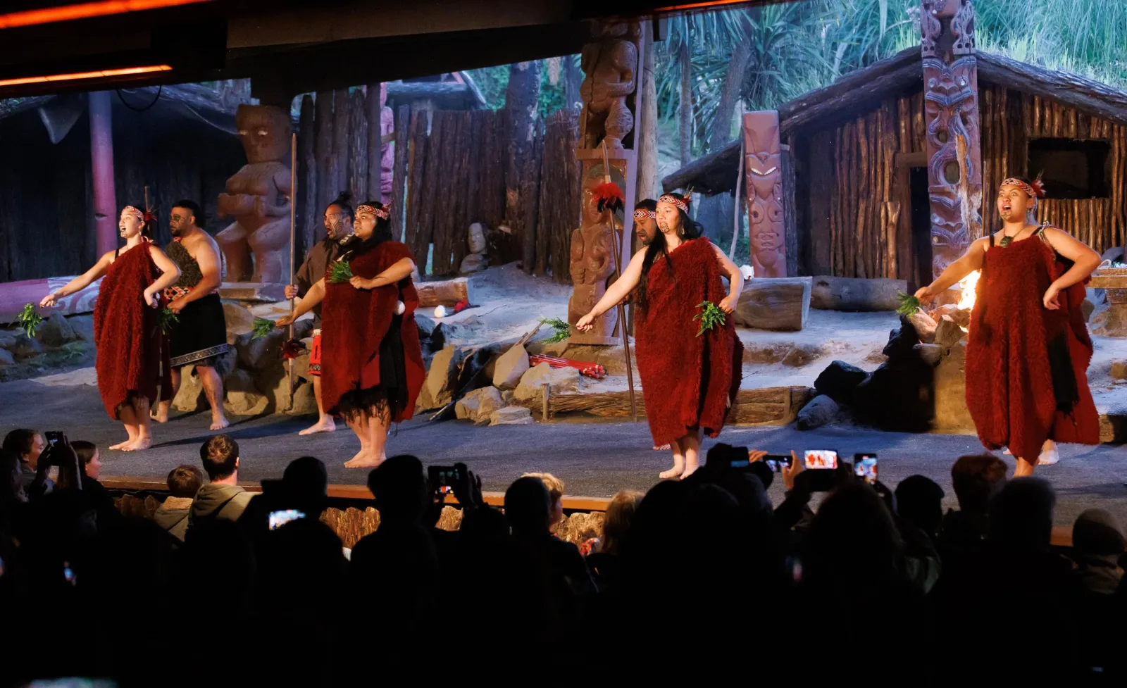 Performers in red korowai on the lamplit stage with the audience silhouetted in front