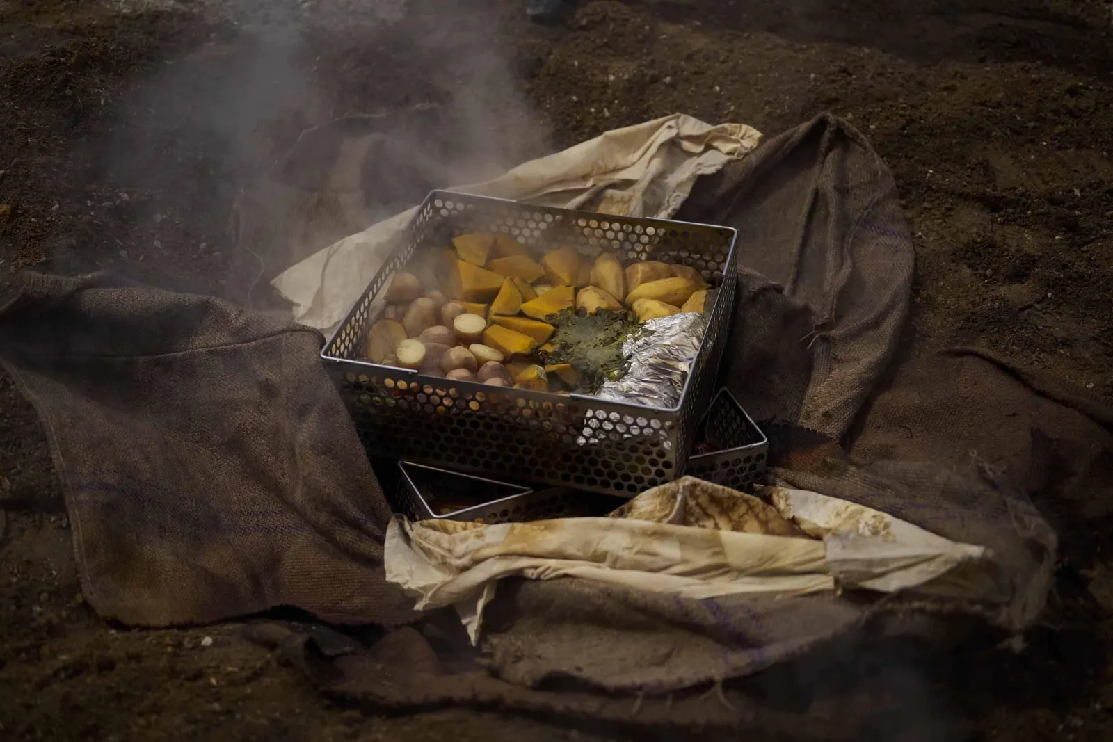 A steaming hāngī basket lifted from the earth oven onto damp sacking