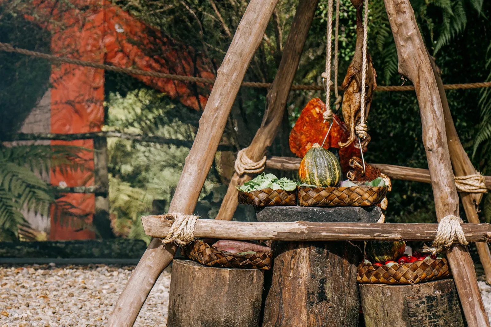 Woven kete of vegetables hanging from a wooden tripod above the cooking fire