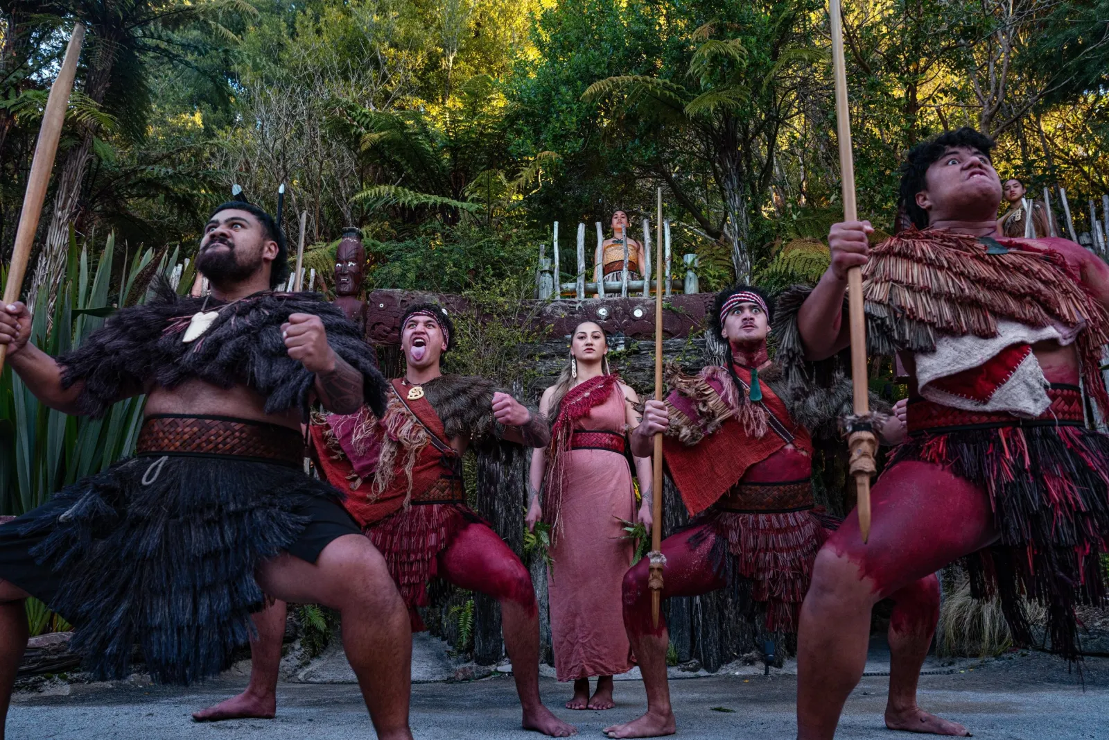 Performers in kākahu raise taiaha during the welcome at dusk
