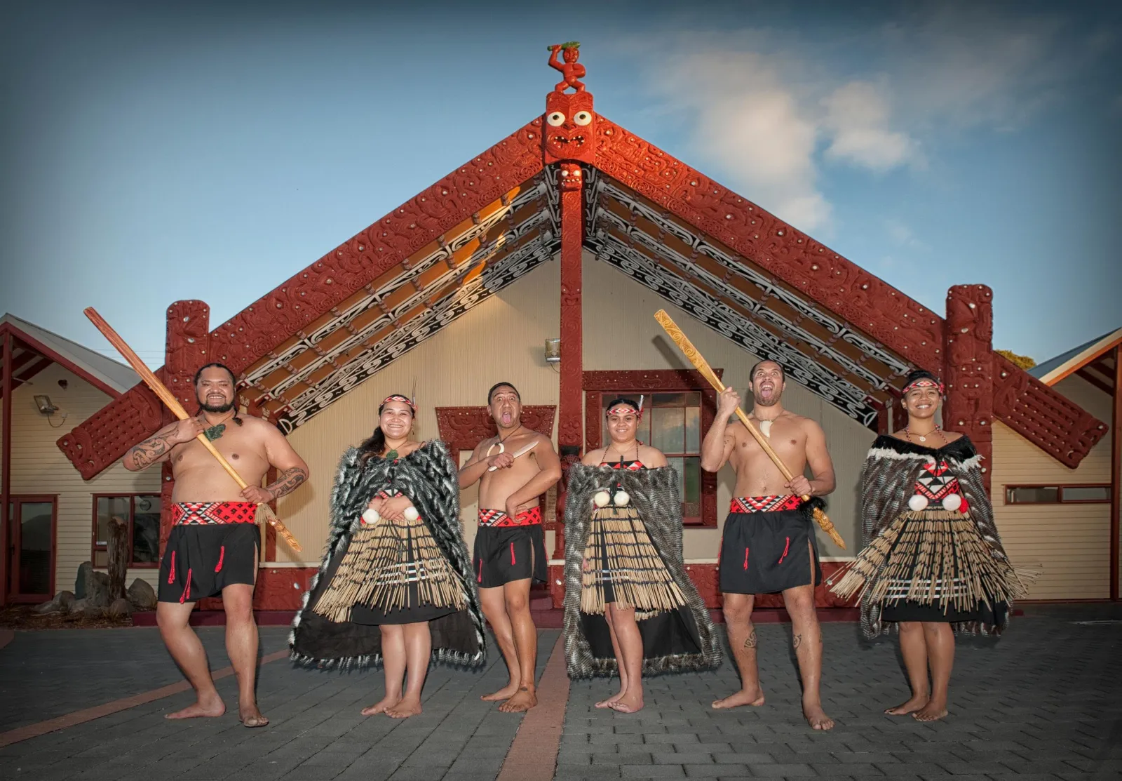 The full performance group in front of the carved wharenui under a blue sky